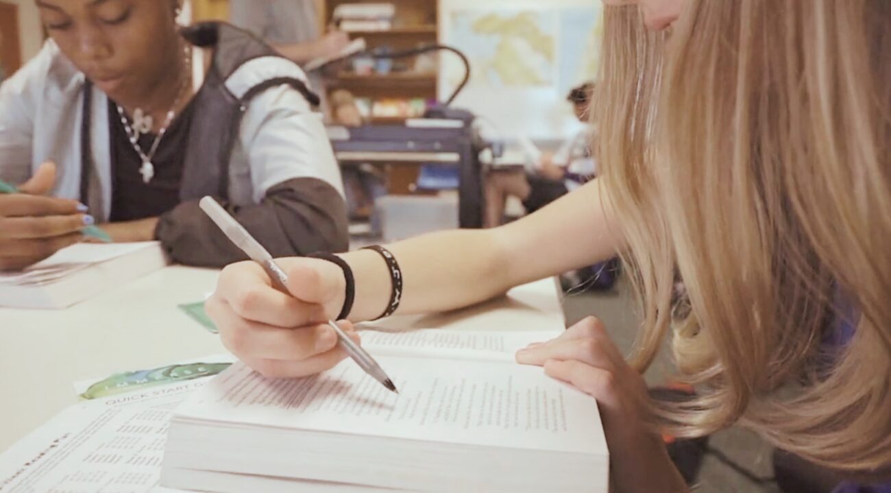 Two students sitting at a desk in a classroom, reading Immerse books