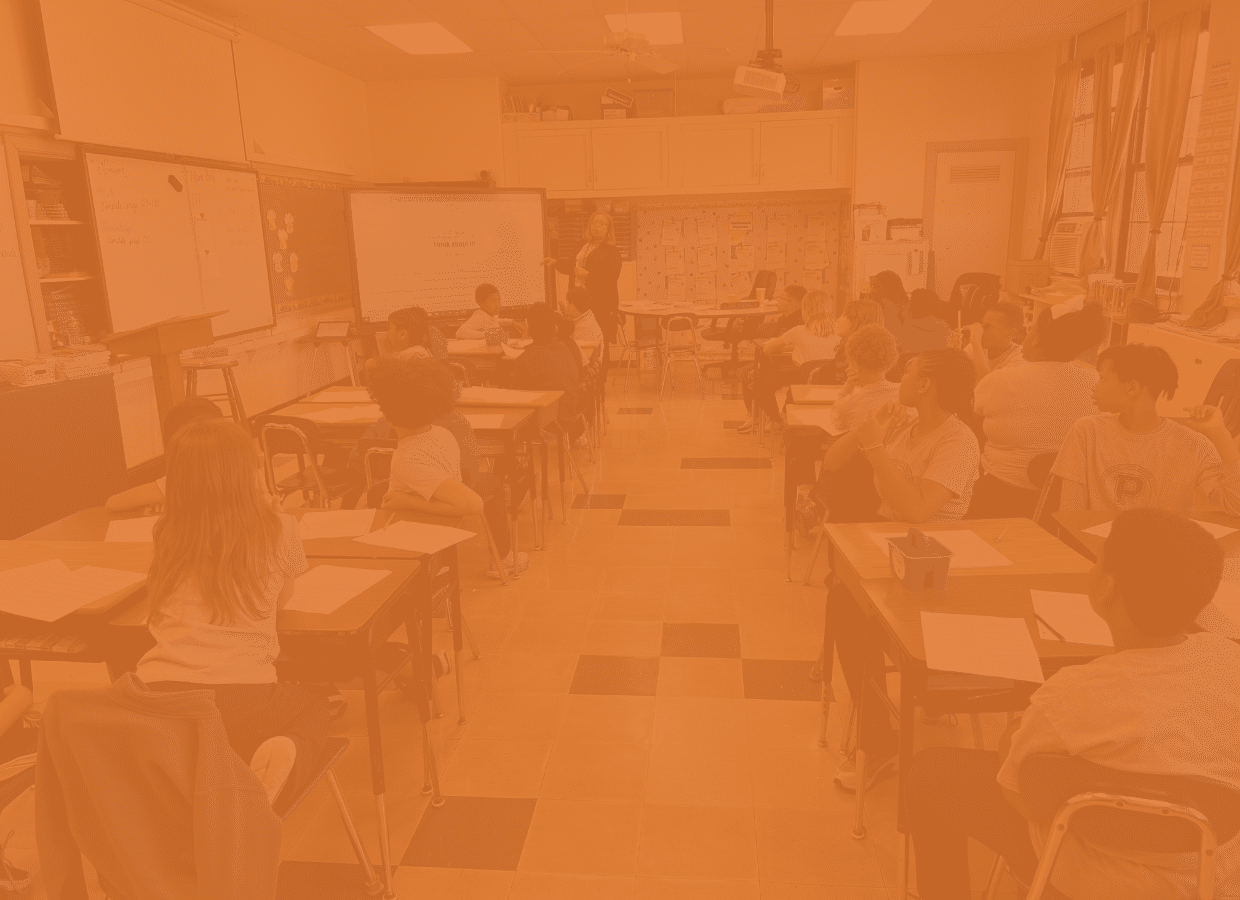 A classroom with students seated at desks facing a teacher presenting at the front near a whiteboard and screen.